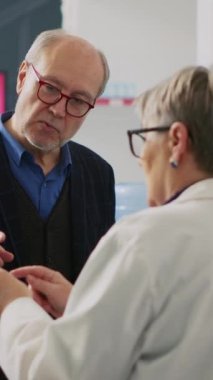 Dikey video: Eczacı üst düzey müşterilere reçete kâğıdı için yardım ediyor, ilaç buluyor ve hastalık tedavisi için ilaveler buluyor. Eczane tezgahında ilaç arayan yaşlı bir adam, yazar kasa..