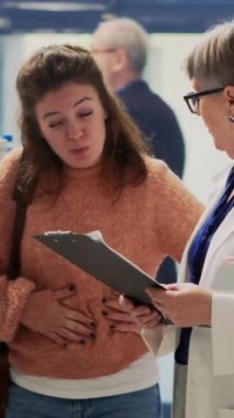 Dikey video: Midesi ağrıyan genç bir müşteri, eczanedeki gastrit hapları hakkında uzmanlara soru soruyor, karın kramplarına karşı ilaç arıyor. Gıda zehirlenmesi ve mide bulantısı çeken kişi..