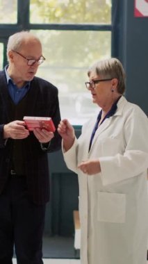 Dikey video: Yaşlı adama kardiyoloji hapları tedavisini açıklayan kadın uzman, broşür okumak için ilaç kutusu tutuyor. Eczanede ilaç ve hastalık tedavisini soran bir müşteri var..