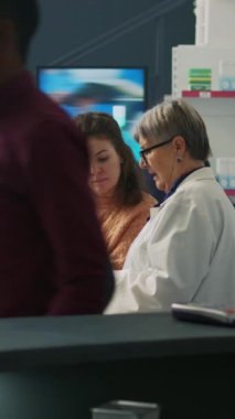 Dikey video: Eczacı müşteriyle ilaveler ve haplar hakkında konuşuyor, eczane raflarındaki ilaç kutularına bakıyor. Uzman, genç bayana tedaviyi ve tıbbi broşürleri açıklıyor..