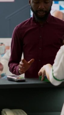 Dikey video: İlaç parasını cep telefonuyla ödeyen erkek müşteri posizyon terminalinde ilaç ve ilaç kutularını alışveriş poşeti şeklinde eczaneden alıyor. Eczanenin kasasında duran adam.