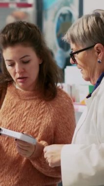 Dikey video: Kadın müşteri vitamin kutusunu kontrol ediyor ve eczacıyla tedavi hakkında konuşuyor, destek paketlerine bakıyor. Genç bir kadın ilaç ve eczacılık hakkında sağlık uzmanına soru soruyor..