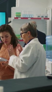 Dikey video: Yaşlı eczacı müşteriye ilaveler ve uyuşturucular hakkında konuşuyor, eczane raflarındaki ilaç kutularına bakıyor. Uzman, tedaviyi ve tıbbi broşürleri...