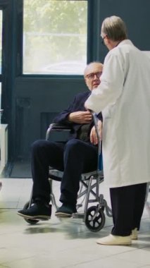 Dikey video: Eczacı yaşlı tekerlekli sandalye kullanıcısına kardiyoloji hapı şişesi veriyor, kronik özürlü bir adam destek istiyor. Eczacılık tedavisi gören engelli bir müşteri