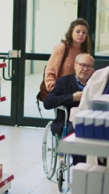 Dikey video: Tekerlekli sandalye kullanıcısı ve kadın bakıcı uzmanlara ilaç almak için reçete kağıdı veriyor. Genç hademe, eczanede kronik bozukluk olan bir yetişkine yardım ediyor.