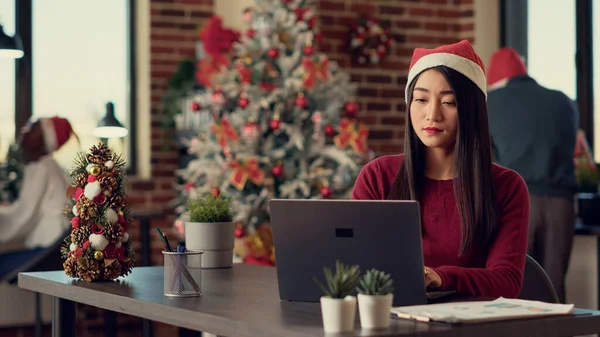 亚洲女人戴着圣诞礼帽 分析笔记本电脑上的数据 在节日办公室做创业报告 圣诞佳节前夕 在带有季节性装饰和装饰品的太空中发送Pc电子邮件 — 图库照片