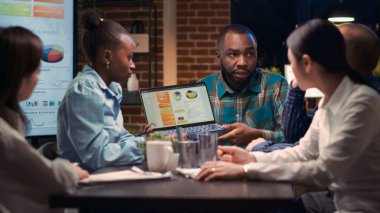 İş toplantısı, Afro-Amerikan analitik uzmanı şirketin gelir raporlarını dizüstü bilgisayardan gösteriyor. İş arkadaşları istatistik sunumunu tartışıyor, çalışanlar analistleri dinliyor.