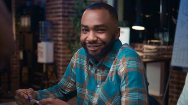 Gülümseyen Afro-Amerikan bir çalışan iş dünyasının portresiyle tanışıyor, yavaş çekim orta ölçekli. Kameraya bakan adam, çeşitli iş arkadaşları beyin fırtınası, planlama stratejisi