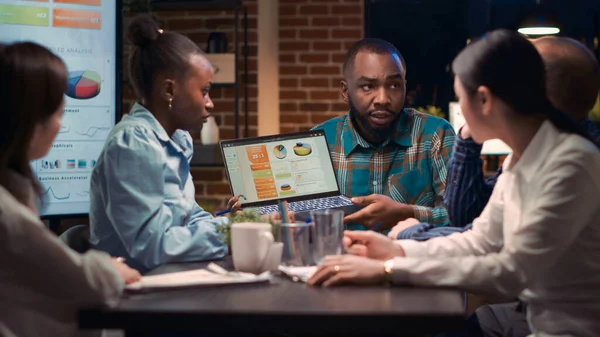 İş toplantısı, Afro-Amerikan analitik uzmanı şirketin gelir raporlarını dizüstü bilgisayardan gösteriyor. İş arkadaşları istatistik sunumunu tartışıyor, çalışanlar analistleri dinliyor.