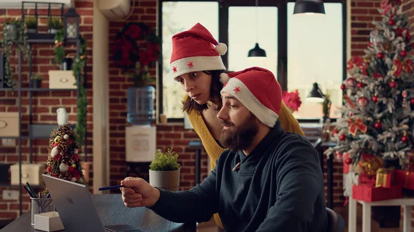 同事们在圣诞前夕的节日办公室里分析笔记本电脑上的报道 戴着圣诞礼帽的男男女女带着圣诞树装饰在太空中进行团队合作 — 图库照片