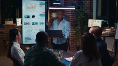 Çalışan finansal raporu sunuyor, dizüstü bilgisayardaki çizelgeleri gösteriyor. Afro-Amerikan kadın istatistik sunumu, çeşitli takımlar analitiği tartışıyor, satış stratejisi planlıyor
