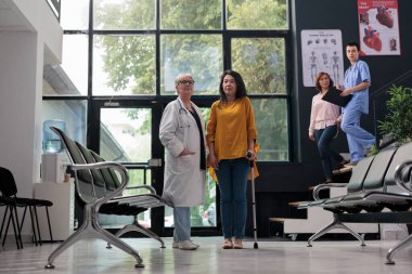 Kıdemli hekim, bacak yaralanması olan bir kadının yürüme iskeleti kullanmasına yardım ediyor. Kırık üstüne fiziksel bozukluklarla uğraşıyor. Asyalı kadın yürüyen yardımla fizik tedavi uyguluyor..