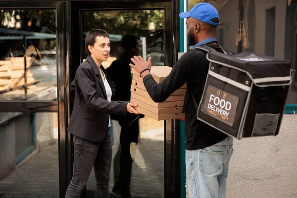 Pizzeria courier giving customer pizza pile, woman taking order. African american man delivering takeaway meal, company employee receiving fastfood packages stack near office building