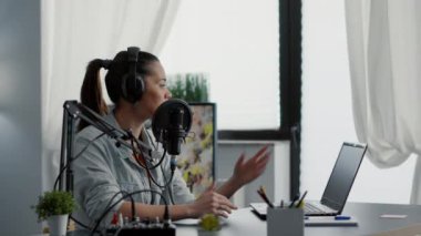 Internet radio show host talking on microphone while checking subject list on laptop. Popular social media content creator speaking to audience while sitting in modern studio.