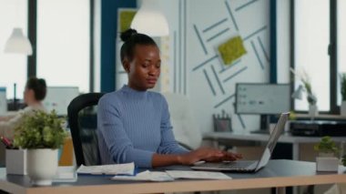 African american startup employee holding business papers with charts looking at laptop screen comparing data. Woman in busy office analyzing marketing statistics and annual turnover.
