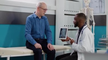 Afrikalı Amerikalı doktor yaşlı adama ait radyografi sonuçlarını analiz ediyor. Reçeteli tedavi için kemik teşhisini inceliyor. Doktor dijital tabletteki x-ray taramasına bakıyor..