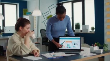 African american woman presenting sales charts using laptop to coworker drinking coffee or tea in morning meeting. Team of colleagues analzying market share looking at portable computer screen.