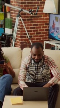Dikey video: Afro-amerikan modern çift evde koltukta dinleniyor, akıllı telefondaki sosyal medya uygulamalarında kayıyorlar. Eğlencenin tadını çıkarmak, birlikte gülmek, oturma odasında eğlenmek..