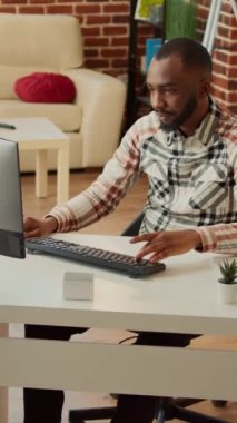 Dikey video: Masada çalışan Afrikalı Amerikalı bir adam, evde kız arkadaşıyla uzaktan kumanda yapıyor. Şirket raporu için finansal veri planlamak için online web uygulaması kullanan genç, e- posta gönder.