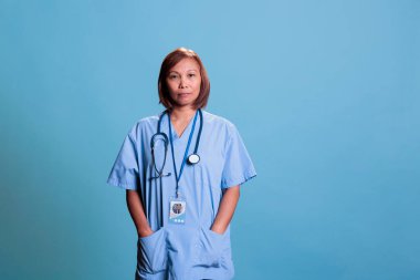 Asyalı asyalı asistanın portresi mavi arka plan üzerinde tıbbi üniforma ve steteskop ile hasta hasta ziyaretine hazır bekliyor. Sağlık hizmeti ve konsept