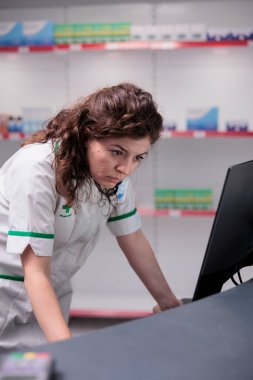 İlaç reçetesine bakan eczane kasiyeri bilgisayar monitöründe ilaç listelerini kontrol ediyor. Eczanede çalışan eczacı, ilaç, vitamin ve takviye malzemeleri satıyor. Sağlık hizmetleri desteği