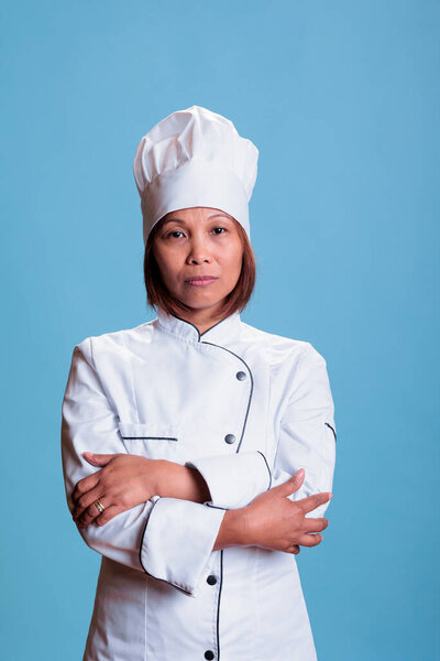 Elderly cheerful chef standing with arm crossed in studio preparing culinary recipe using healthy ingredients on blue background. Woman cook with white uniform and apron cooking meal dish.