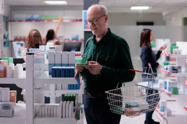 薬局で病気の薬を探す薬棚を見て買い物かごを持っている高齢者 ドラッグストアで丸薬のラベルを読んでいる陽気な男 医薬品 — ストック写真