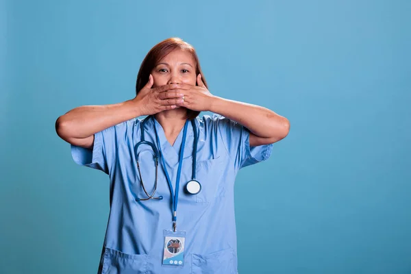 Physician nurse wearing medical uniform and stethoscope covering mouth with hands, three wise monkeys concept. Specialist assistant working at health care expertise, speak no evil,