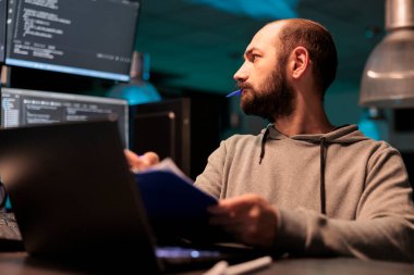 Web geliştiricisi, kaynak kodu kağıtlar ve bilgisayarla analiz ediyor, notlar alıyor ve web geliştirme üzerinde çalışıyor. Dosyaları çevrimiçi sunucu verileriyle karşılaştıran erkek programcı.