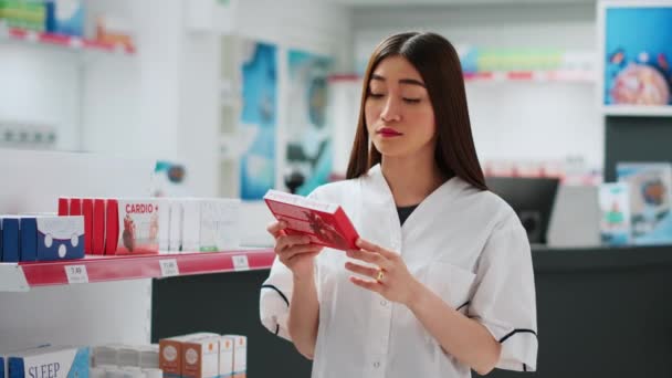 医薬品店で働くアジア人社員は 心臓病の薬を見ています 薬局の小売店 医療での治療やサプリメントを分析します 手持ち撮影 — ストック動画