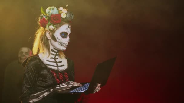 Santa Muerte女士在工作室的笔记本电脑上工作 在无线个人电脑上上网查阅 卡特里娜卡特里娜的模特穿着服装 头盖骨 在音乐工作室里化妆 手持射击 — 图库视频影像