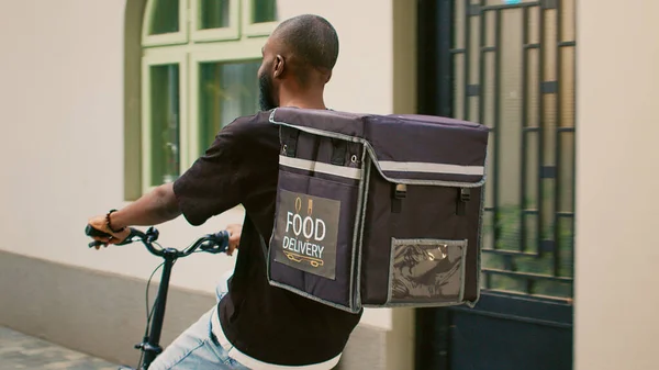 Takeaway carrier going to office building entrance, young man working as food delivery courier on bicycle. African american fastfood employee with thermal bag riding bike on street.