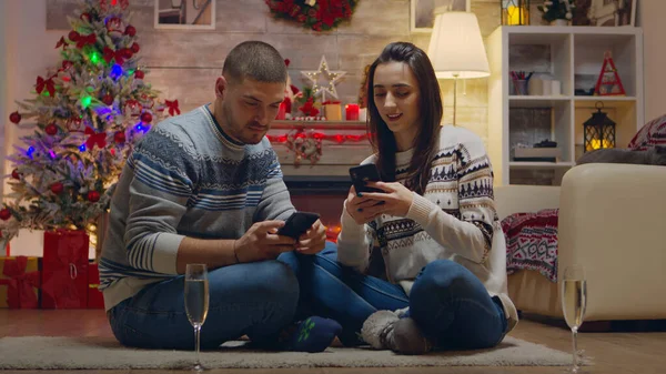 情侣们在壁炉前庆祝圣诞节 用智能手机寻找礼物 香槟杯 — 图库照片