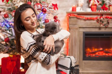 Neşeli genç kadın Noel 'i kutlamak için şöminenin önünde kedisine bakıyor..