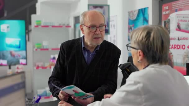 老年人在药店柜台与药剂师交谈 在药品传单上询问药物的情况 年长顾客谈论在药店货架上 药盒上的治疗 — 图库视频影像