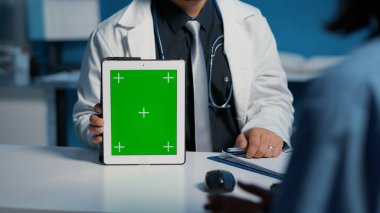 Pratisyen doktor elinde tablet bilgisayarla yeşil ekran kroma tuşuna işaret ederek tıbbi uzmanlığı açıklayan bir görüntü oluşturuyor. Sağlık ve hemşire muayene randevusu sırasında saatlerce çalışıyorlar.