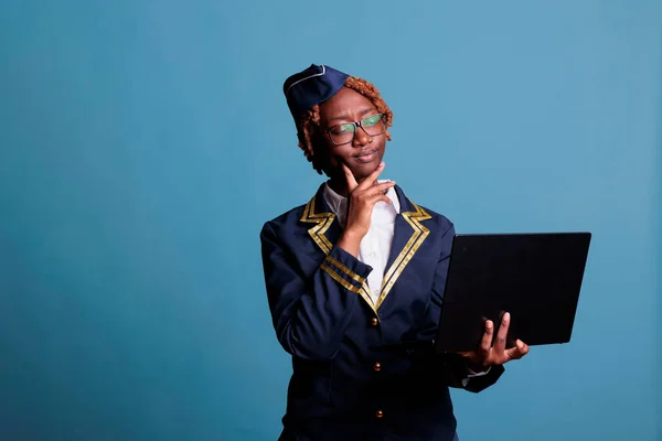 Thoughtful female african american flight attendant in uniform using laptop in studio shot. Stewardess looking doubtfully at new directives received by mail against a blue background.