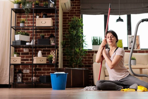 Woman sitting on floor holding mop disappointed doing household chores, looking off into distance while imagining being somewhere else. Housewife tired of being in charge of cleaning and tidying up.