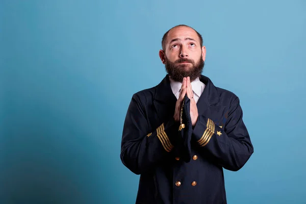 Airline pilot in aviation uniform praying to gods with closed eyes, standing with folded hands gesture. Religious middle aged plane aviator pleasing, aviator worshiping before flight, praising