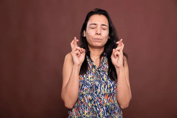 Indian woman crossing fingers, pleading for success, praying for luck, making wish. Girl getting lucky, making wish, standing with hopeful facial expression, studio medium shot on brown background