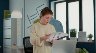 Woman in busy office holding clipboard with sales charts in video call conference on laptop talking about company turnover at desk. Business employee discussing profit with manager over internet.