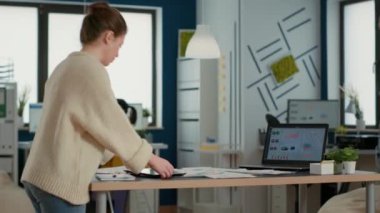 Woman holding digital tablet zooming in on sales pie chart and comparing data with statistics on laptop screen and papers on desk. Startup employee working with key performance indicators in office.