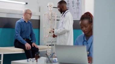 Afrika kökenli Amerikalı osteopat, sağlık kontrolündeki yaşlı hastaya kemik hastalığını ve teşhisi açıklamak için insan iskeletini işaret ediyor. Osteopati muayenesinde omuriliği gösteren anatomi uzmanı..