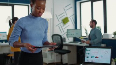 Portrait of african american woman standing in busy office picking up tablet with business erp software and smiling looking at touchscreen. Employee holding digital device and interacting with screen.
