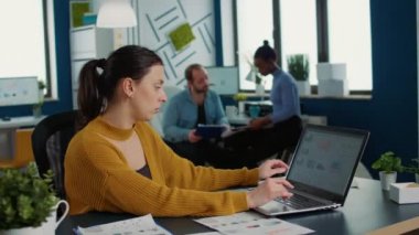 Woman comparing statistics with sales charts on laptop and taking a break to drink coffee or tea sitting at desk. Casual startup employee working relaxed in busy finance department analyzing