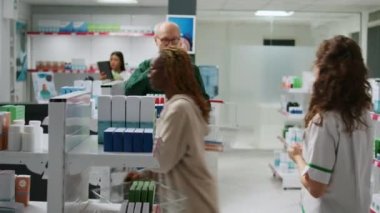 Kadın eczacı, çok ırklı bir grup müşterinin eczaneden ilaç ve vitamin almasına yardım ederek raflardaki haplardan bahsediyor. İlaç ürünleri ve ilaçlara yardım etme.