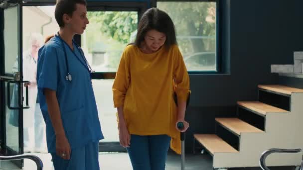 アジアの女性が棒で歩くのを助け 身体障害に苦しみ 杖を使うのを助ける医療助手 施設の足の損傷から回復するためのリハビリテーション療法に参加 — ストック動画