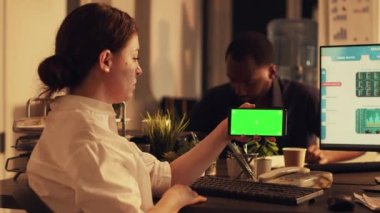 Kadın yönetici elinde yeşil ekran ekranlı akıllı telefonu tutuyor, cep telefonundaki boş krom anahtar geçmişini analiz ediyor. Bilgisayar üzerinde münferit model şablonu ve borsa takası kullanılıyor.