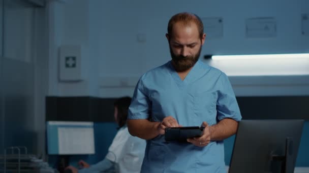 夜間勤務で医療の専門知識を入力しながら 患者診断を確認するタブレット型コンピュータを保持する医師看護師 保健医療で働く青の制服のアシスタントマン — ストック動画