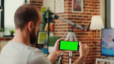 Erkek çalışan cep telefonunda yatay yeşil ekran görüntüsünü kontrol ediyor, izole krom anahtar şablonu olan akıllı telefona bakıyor. Boş kopyalama arkaplanı ile çalışmak.